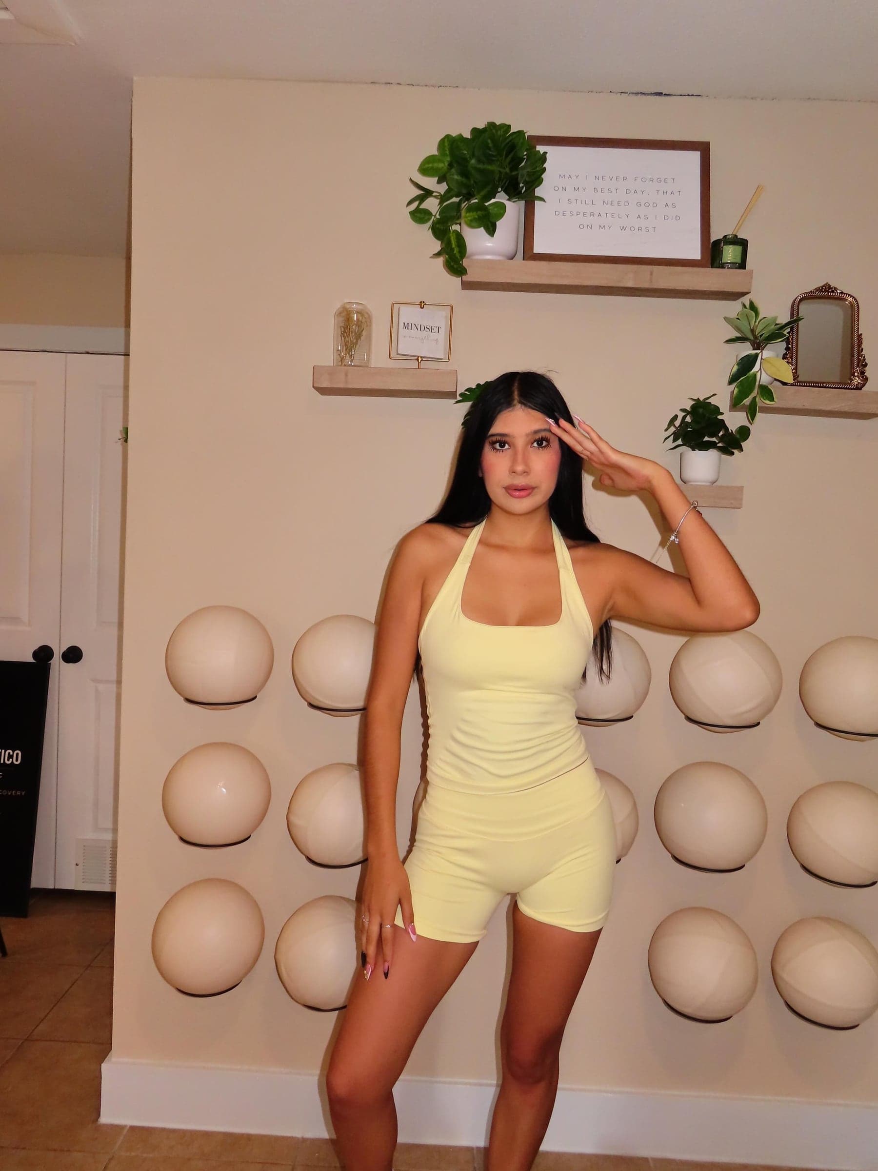 A smiling woman in a yellow set standing in front of the studio's wall of Pilates balls